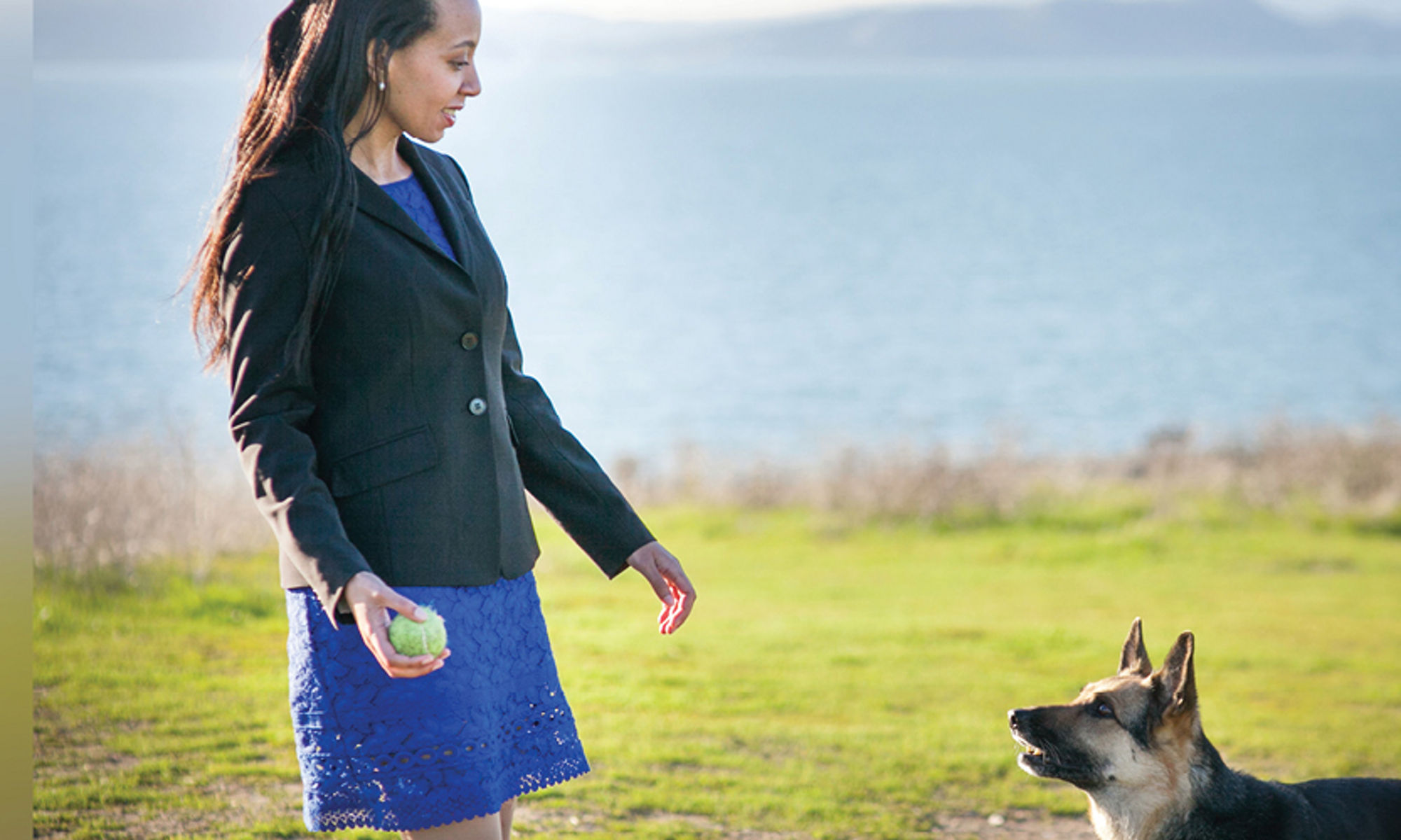 This photo is of Haben standing in a royal blue dress with a black blazer and with a light-green tennis ball in her hand. Her seeing-eye dog sees the ball and anticipates she will play by throwing it. It is a sunny day. They are standing on grass that is green with brown dirt patches. In the background, a body of water is blurred in the top third of the photo.