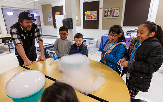 Group of children and a teacher around a table watching a bowl of smoking dry ice.