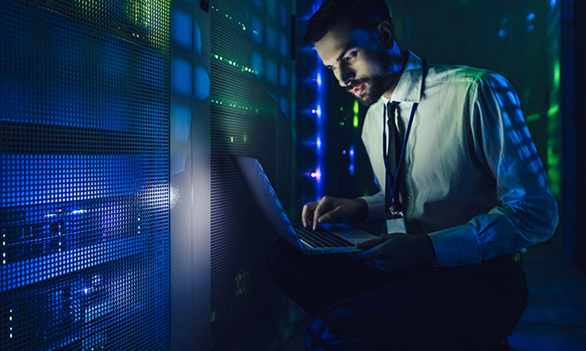 Man in a dimly lit data center, holding a laptop