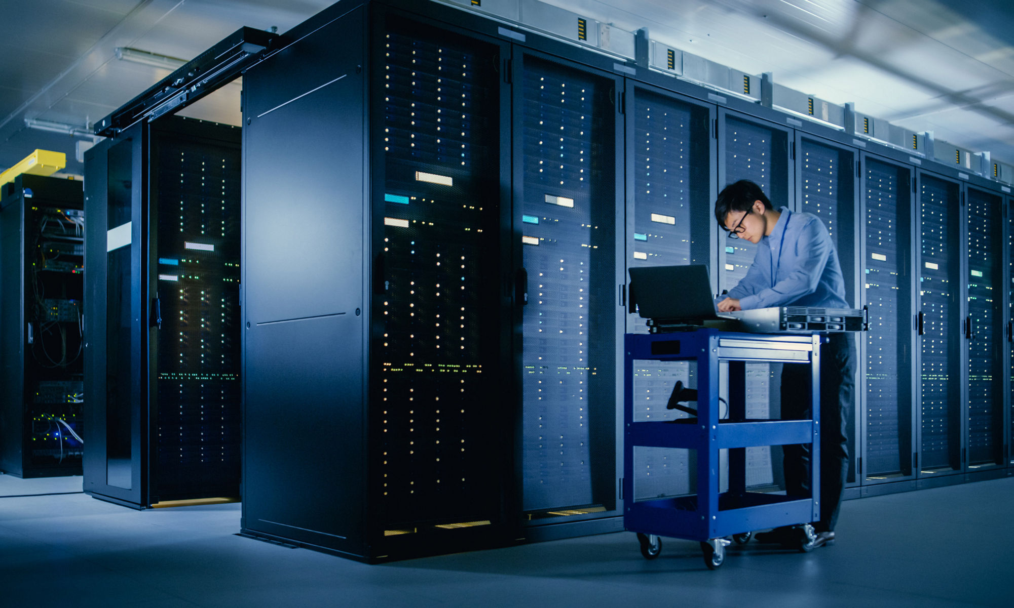 In the Modern Data Center: IT Engineer Doing Maintenance and Diagnostics. Pushes Cart alongside Server Racks, Ready to Install New Hardware for Planned System Update, Equipment Replacement.