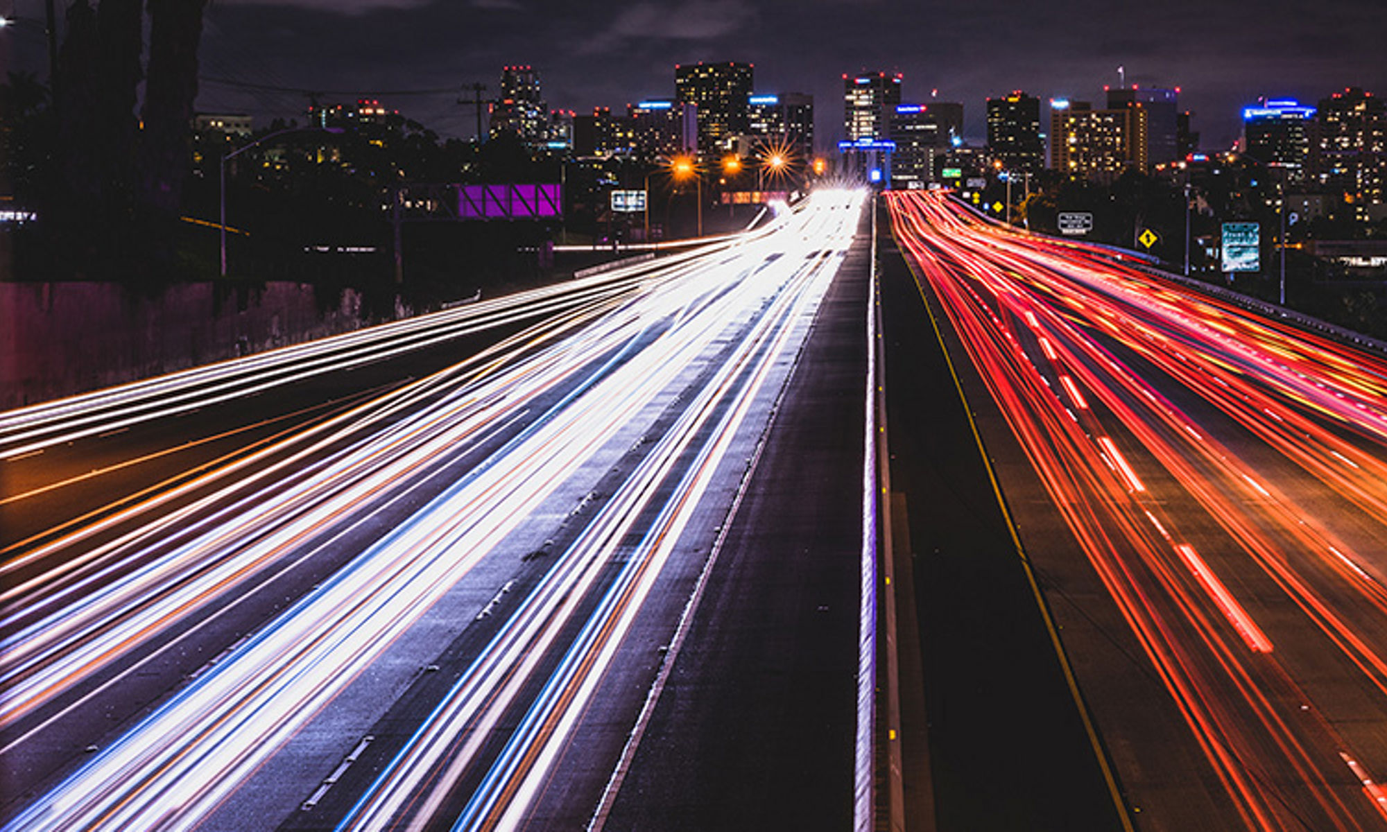 Car headlights and taillights streaking by on a freeway at night with a city in the background