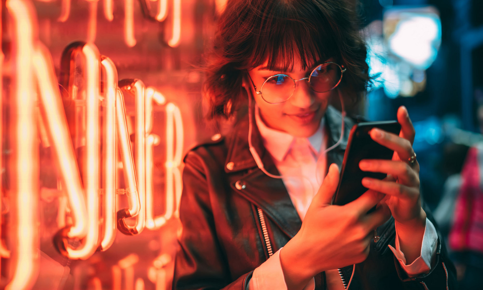 Woman standing in front of a neon sign looking at her phone, with headphones on, wearing a leather jacket.