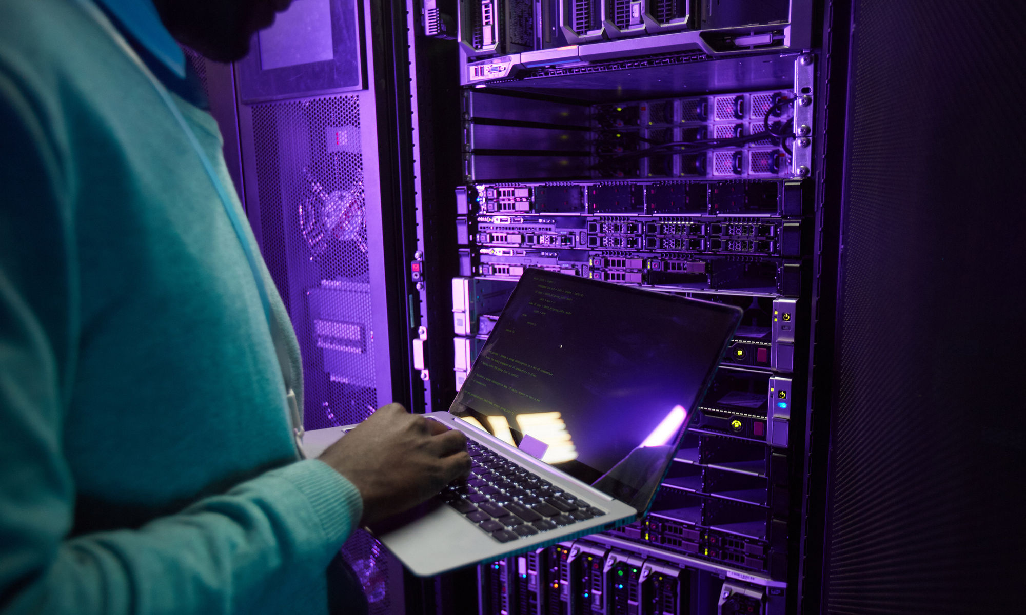 Cropped shot of African American data engineer holding laptop while working with supercomputer in server room lit by blue light, copy space