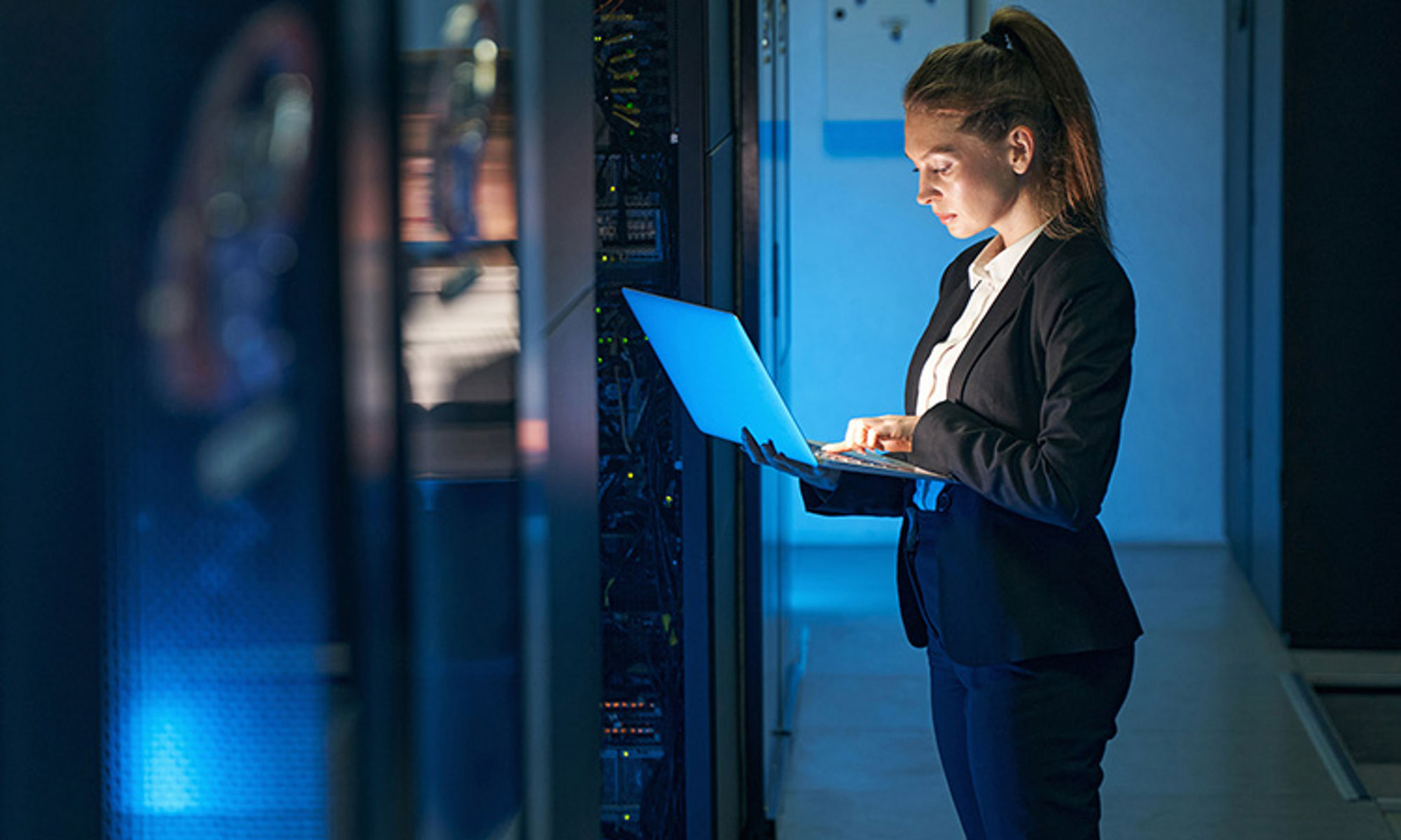 Female worker in dimly lit server room, holding an open laptop