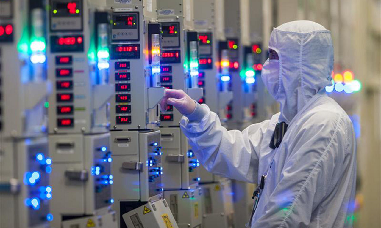 A person in a cleanroom suit inspecting equipment with digital displays and indicator lights in a semiconductor manufacturing facility.