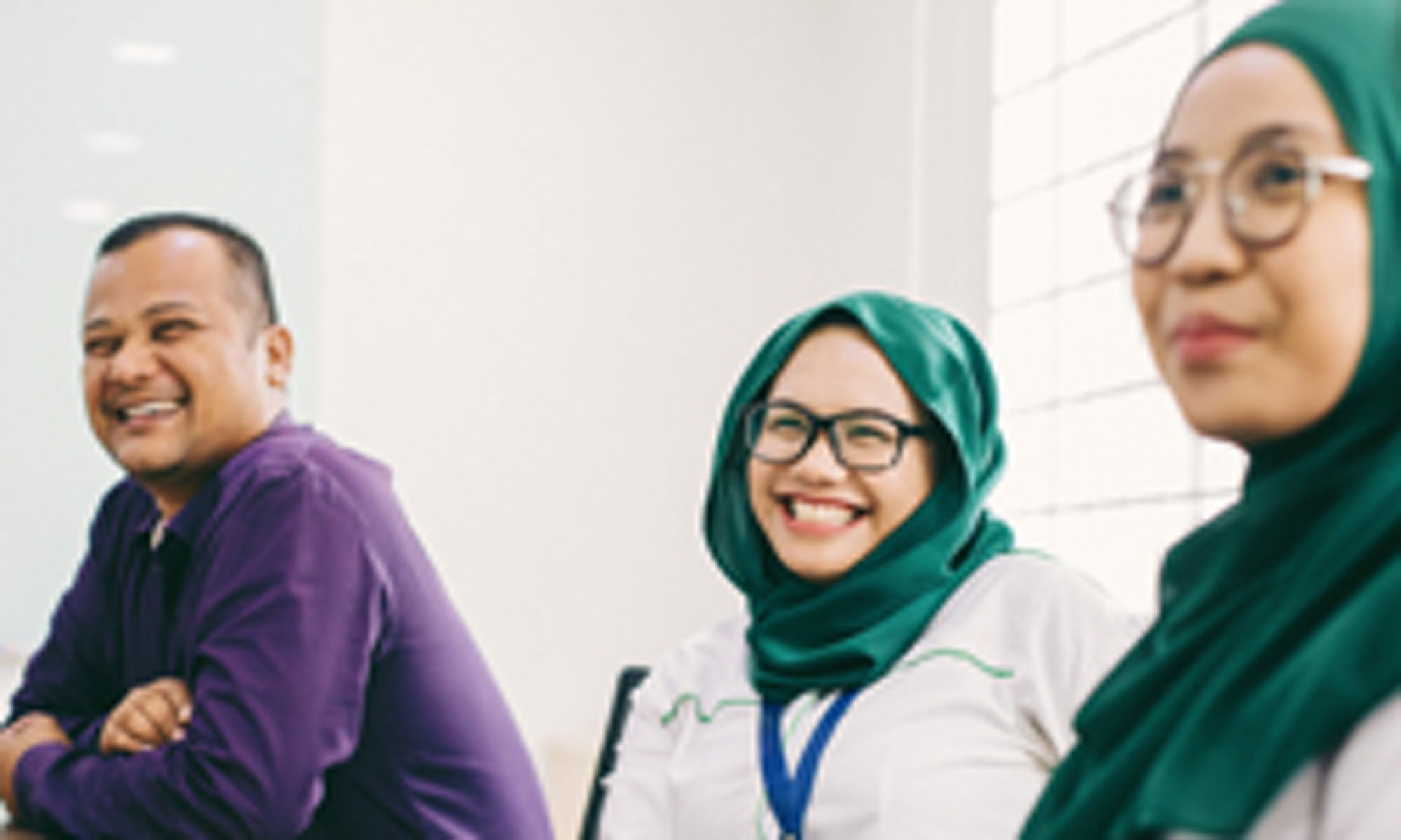 group of people with a man sitting wit folded hands and Two women wearing hijab look happy 