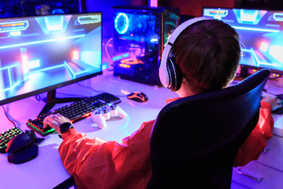 ヘッドフォンをつけた赤いパーカーの人物がカラフルな画面のマルチモニターの前でビデオゲームをプレイしている