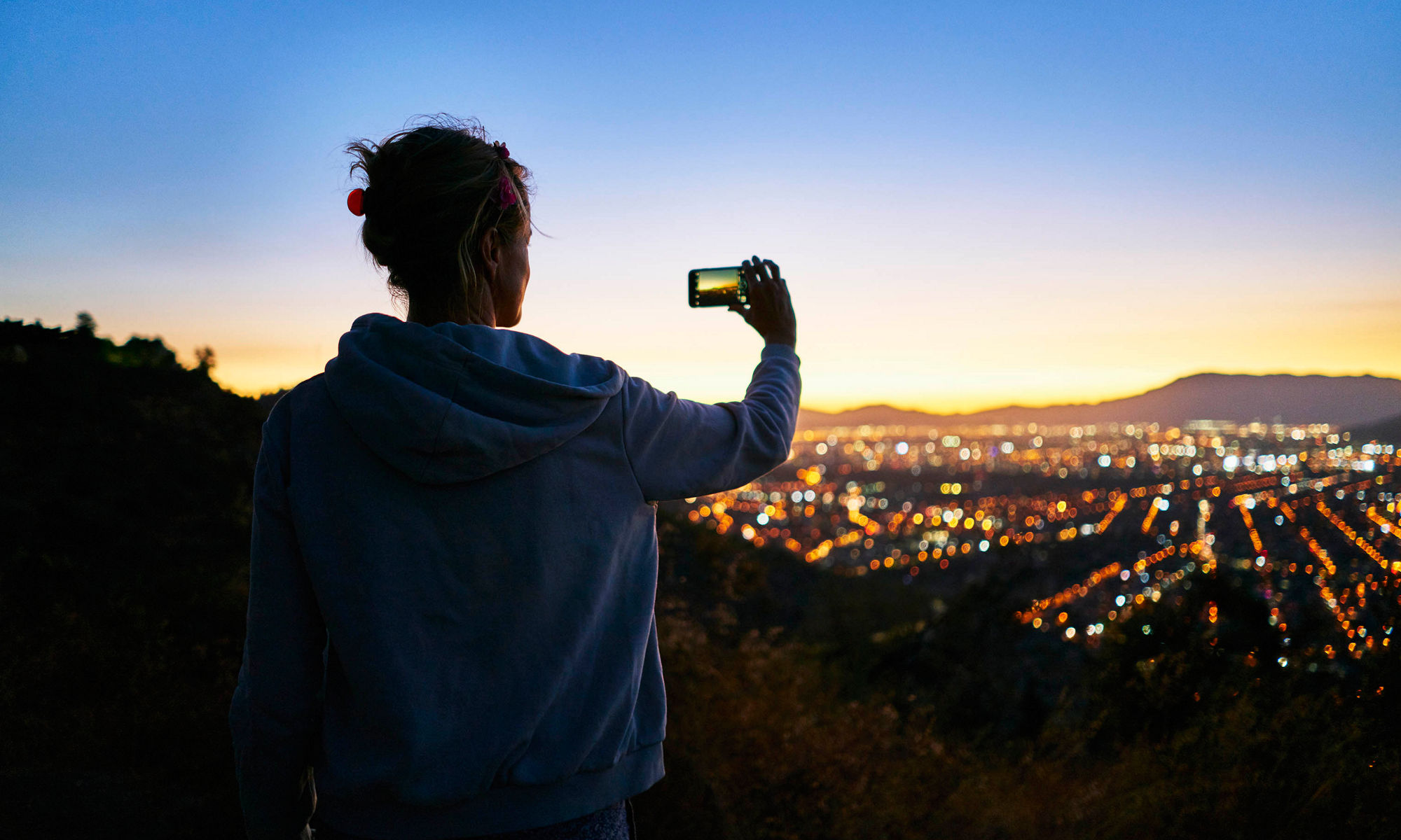 Person's hand holding up a mobile device to take a picture of a city scape blurred in the background