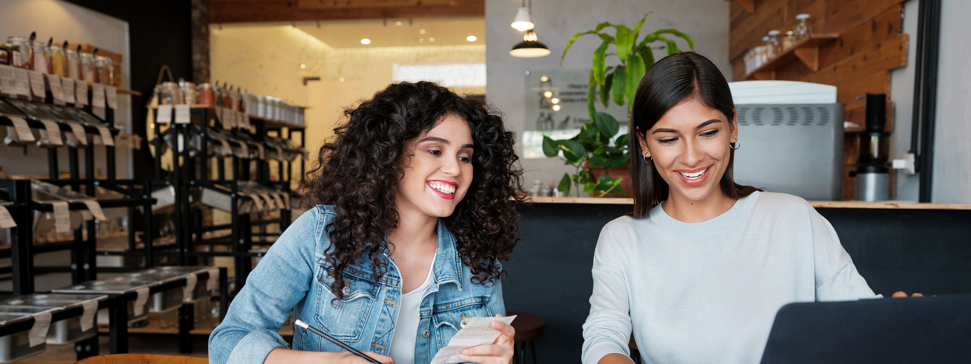 Two young latin women sitting at a table in an eco-friendly store, using a laptop and smiling.