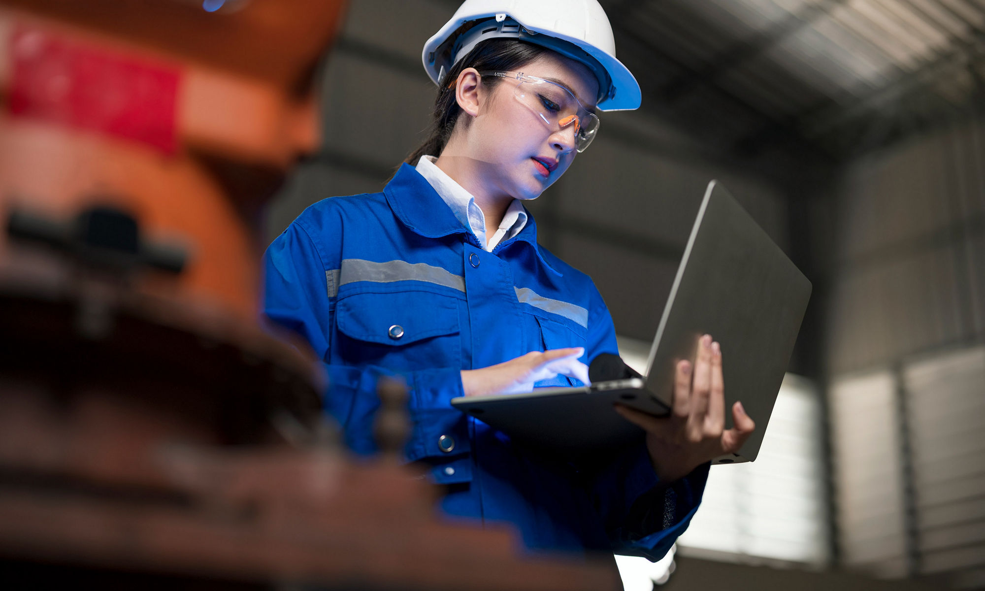 Standing woman wearing a hard hat and blue coveralls working on a laptop