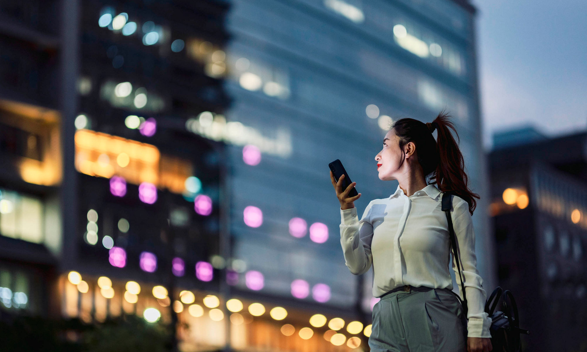 Evening view of city scape over the shoulder of a dark haired woman taking a picture with a mobile device 