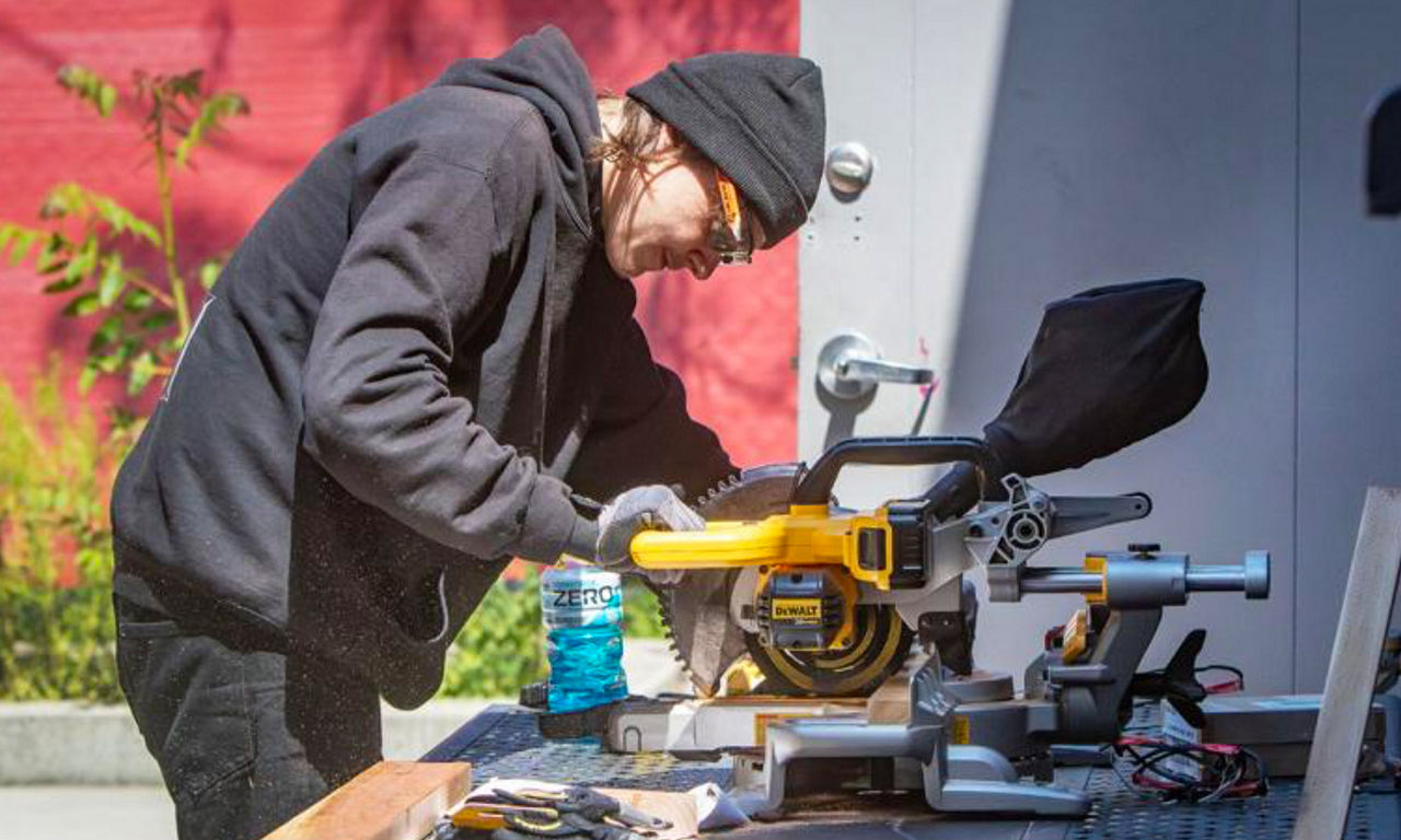 Person in black hoodie using a yellow miter saw on a workbench with various tools