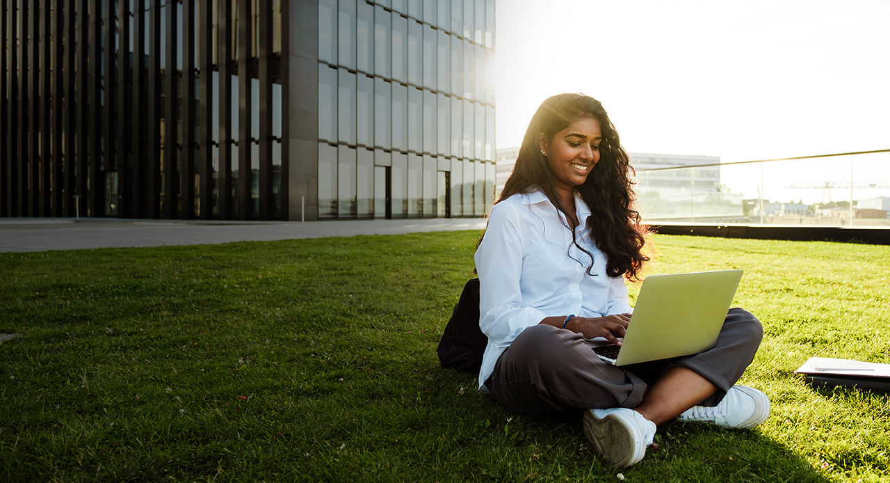 Woman student sitting on grass in front of a building, typing on a laptop