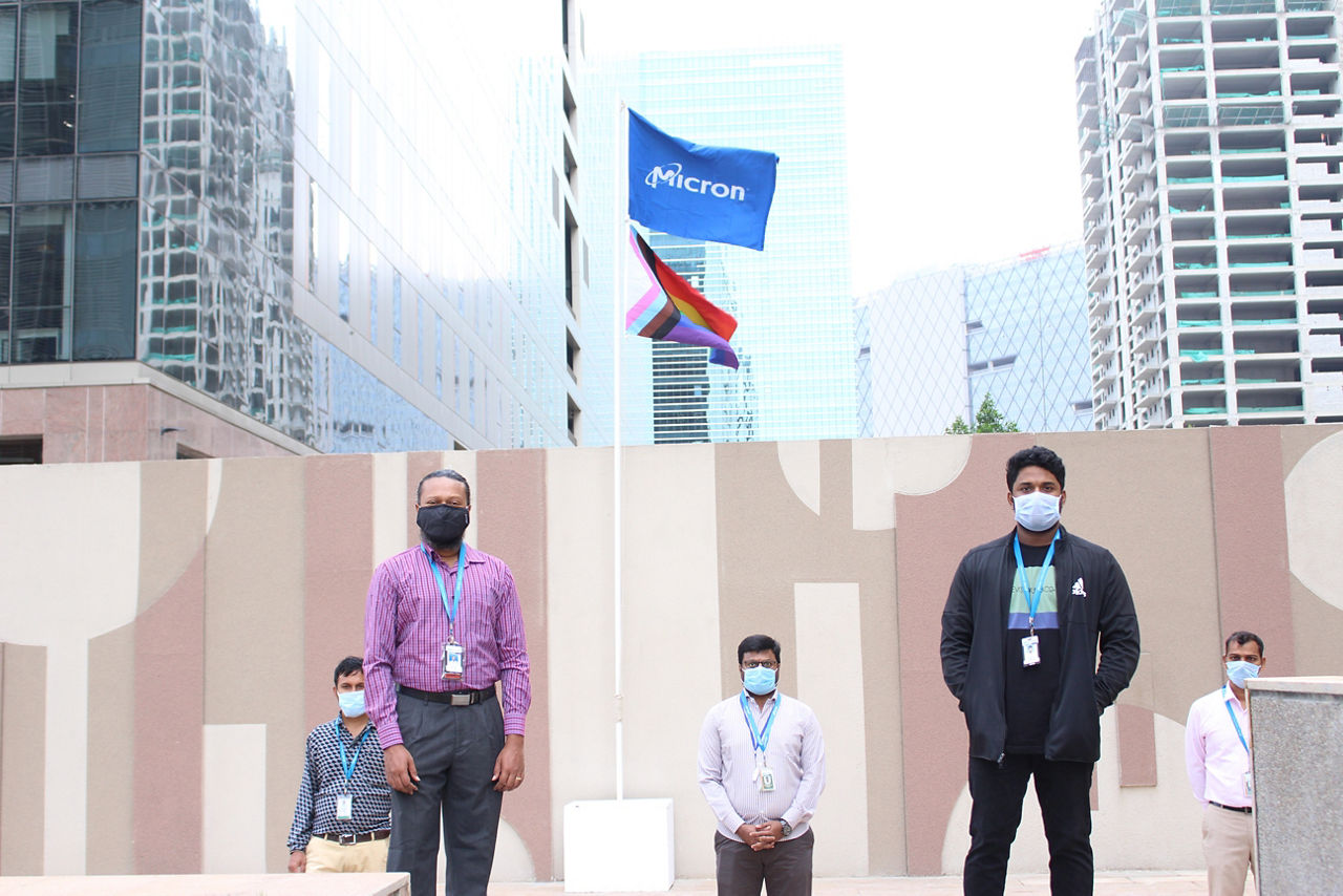 Group of men wearing masks standing couple of feet apart Infront of micron flag