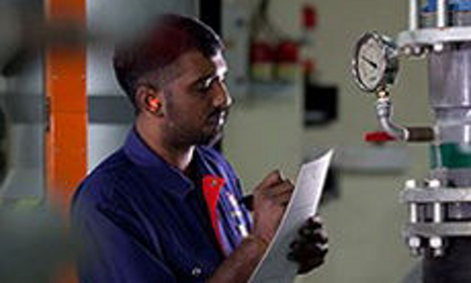 A man writing on paper while standing looking at a pressure guage