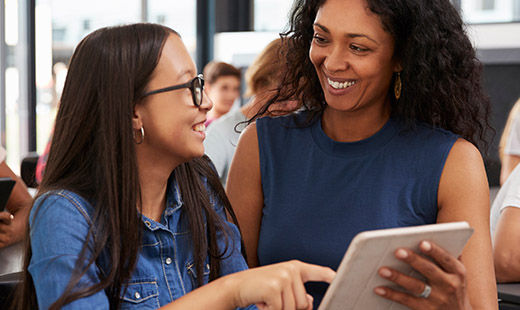 A female teacher and female student smiling to each other, as they discuss something looking at a touch-screen tablet