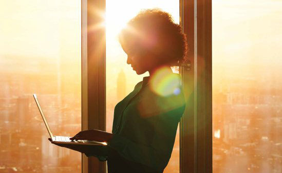 A standing woman of color interacting with a laptop while in fron tof a window with a bright sun shining