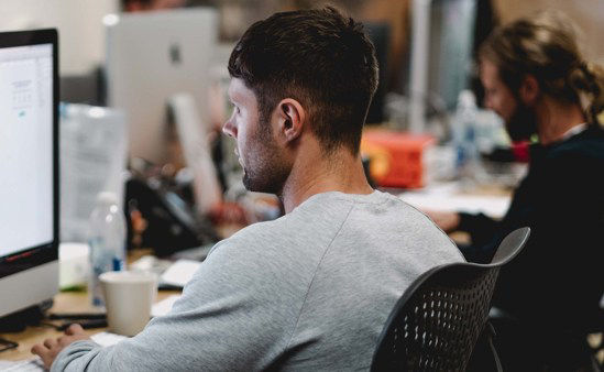 A seated young man reading a computer screen in a work environment