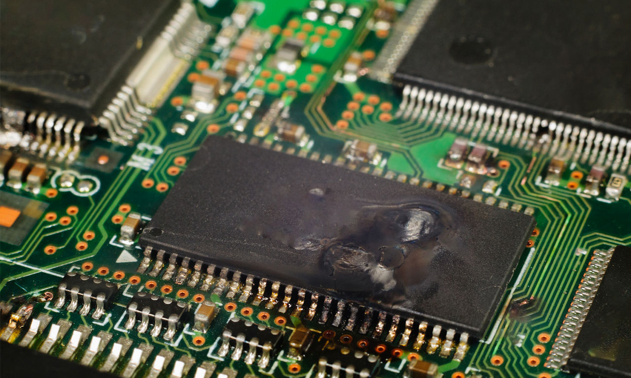 A close-up of a green circuit board with one black microchip in the center appears damaged with visible scorch marks