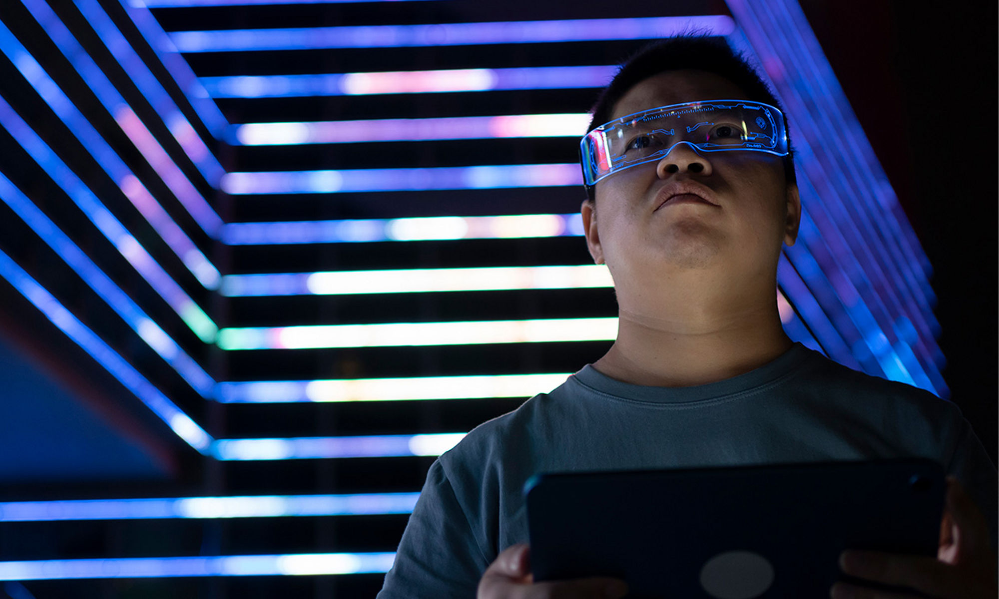 Man wearing goggles holding laptop at night in front of a lighted skyscraper