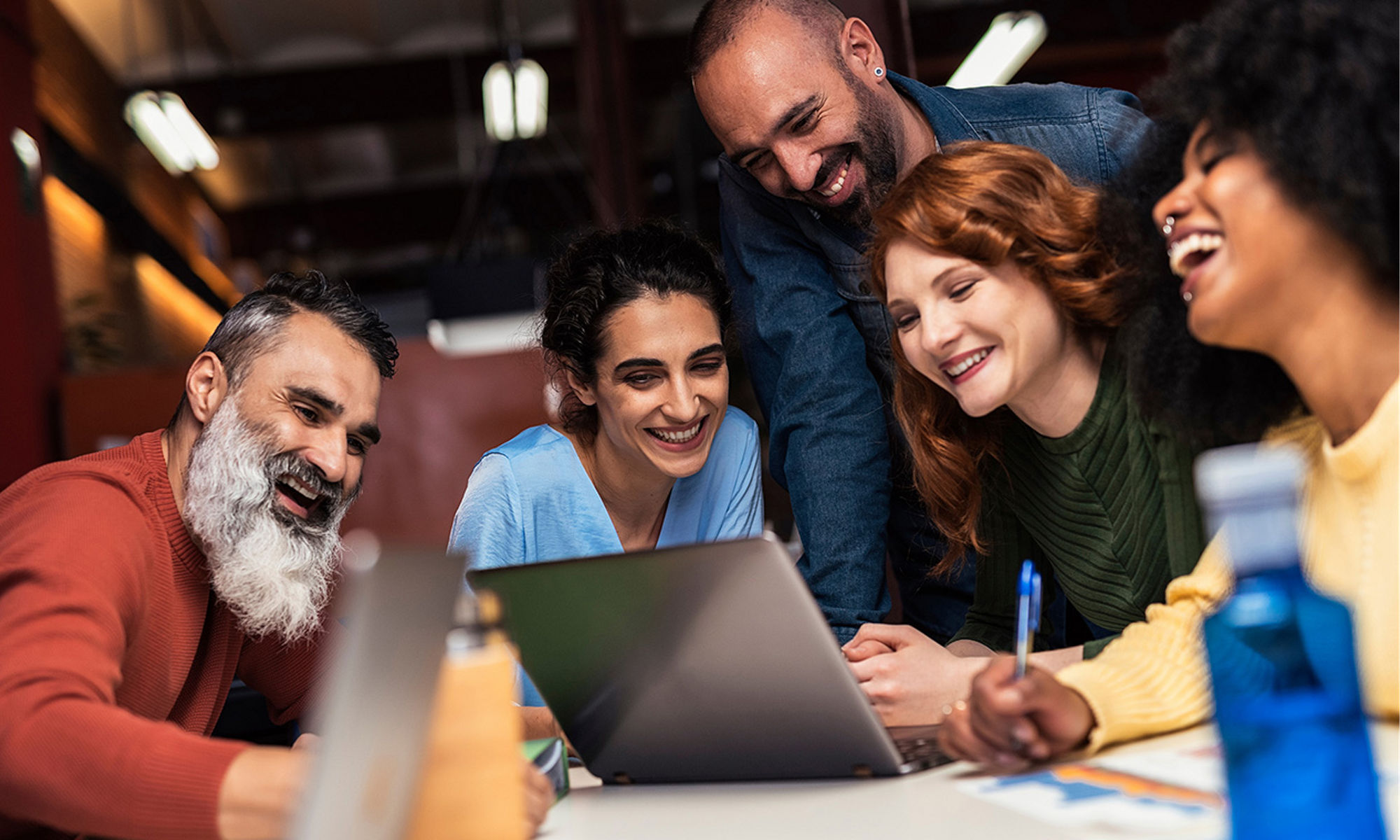 A group of diverse, smiling and laughing people, sitting around a laptop.