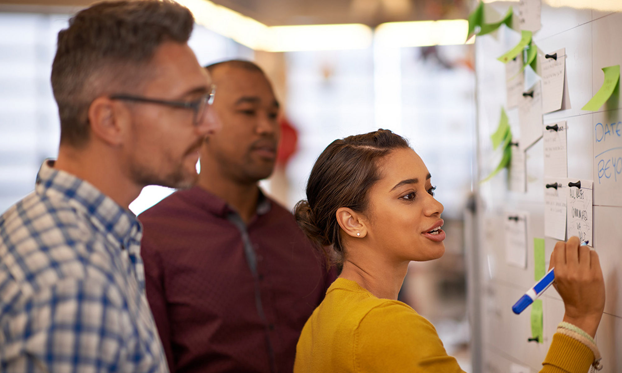 A woman writing on a whiteboard wall with two male co-workers looking over her shoulder.