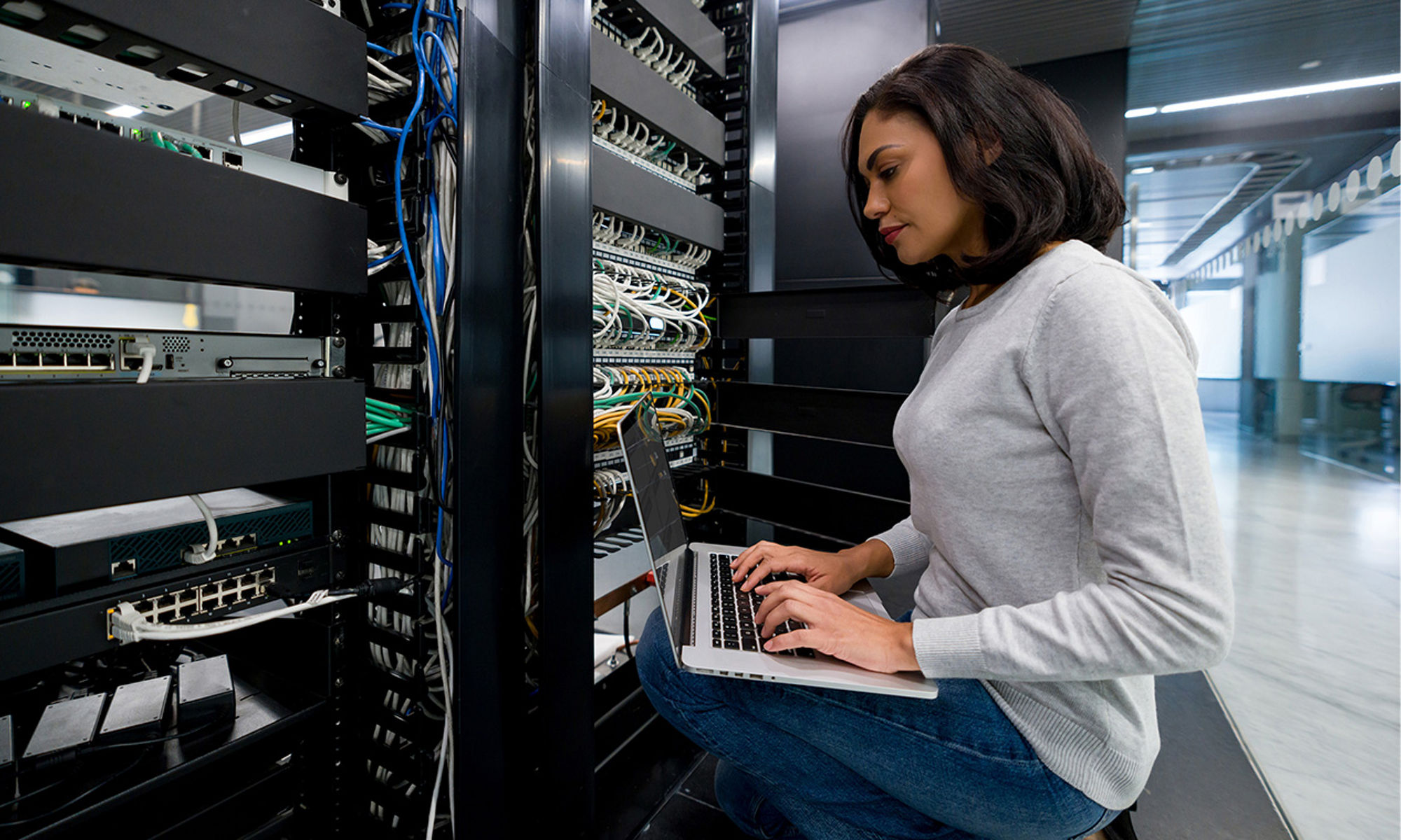 Woman kneeling in front of a server, working on a laptop.