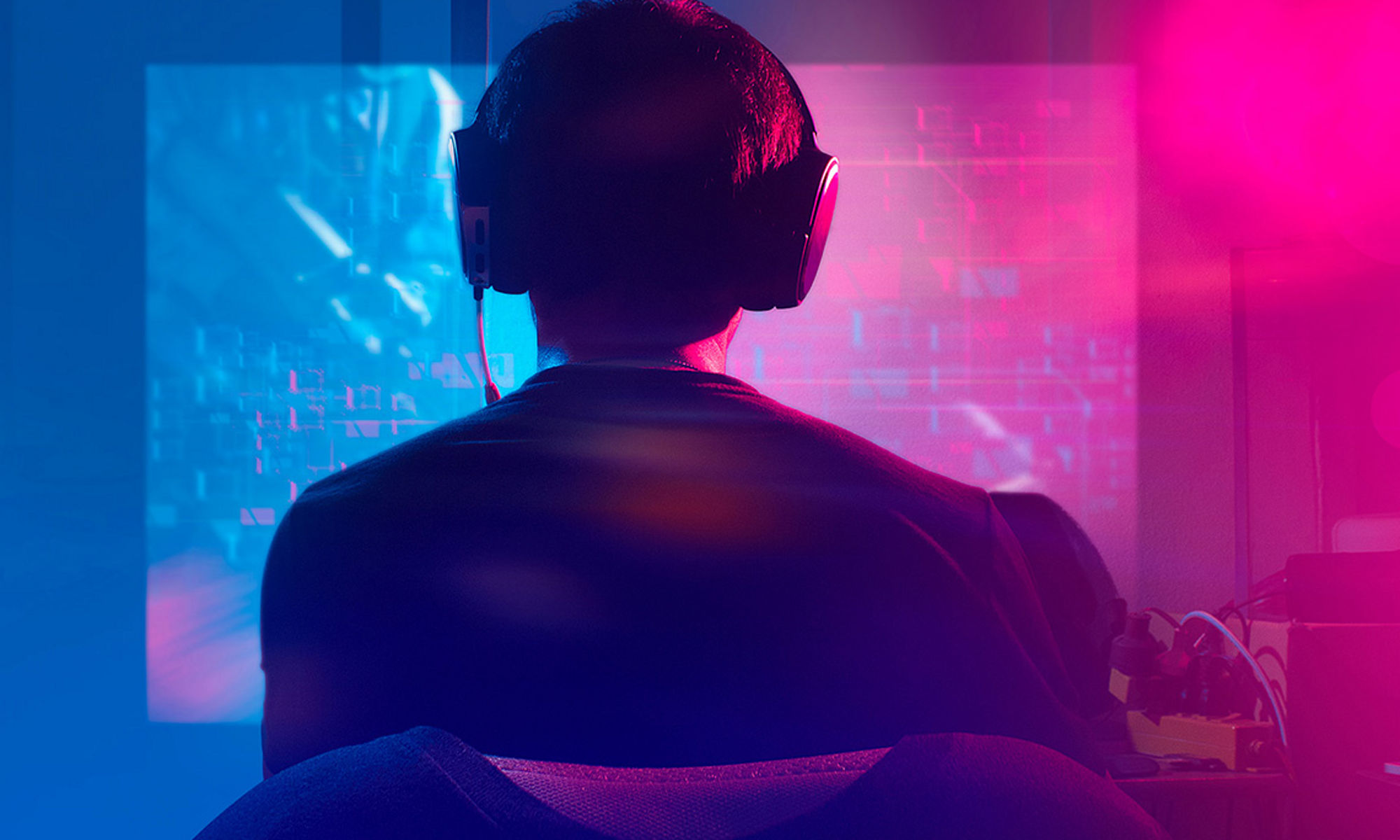 View of back of man while he sits in front of computer with headphones on in pink hues