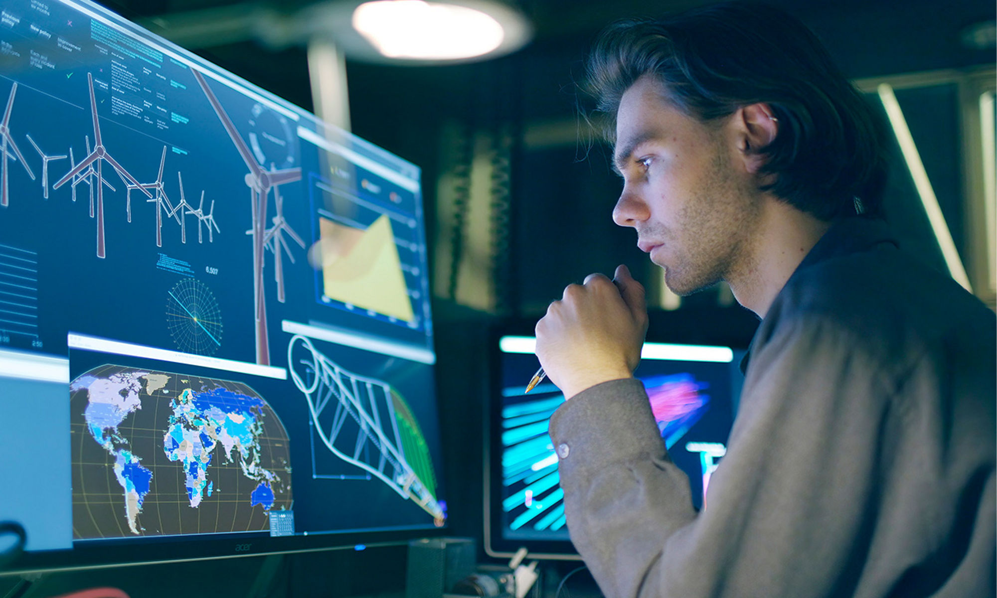 Stock image of a young, long-haired man, working at night in front of a large computer screen showing designs for a wind turbine