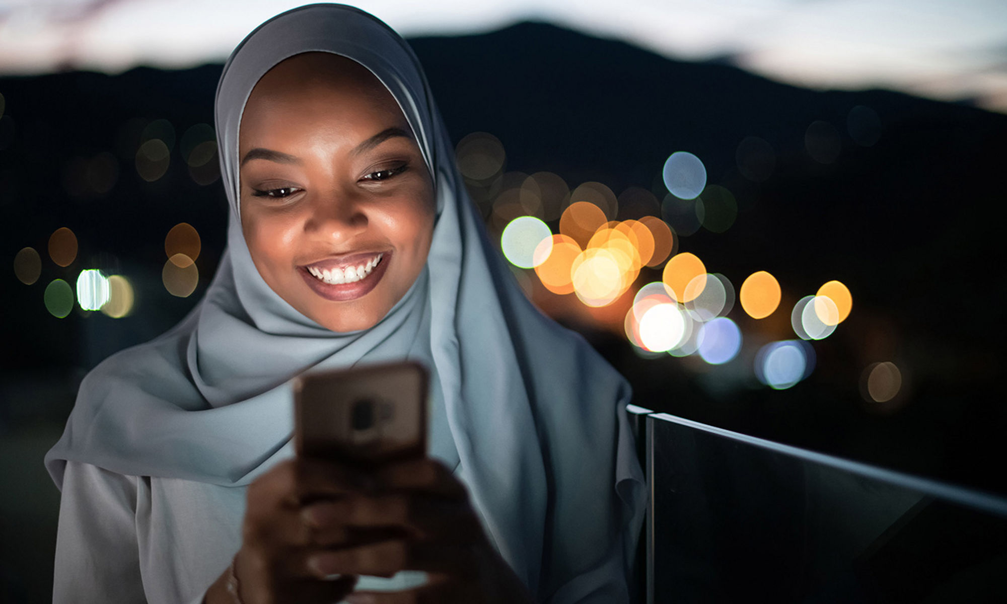 Young Muslim woman outside looking at her mobile phone with blurred sunset sky and city lights behind her