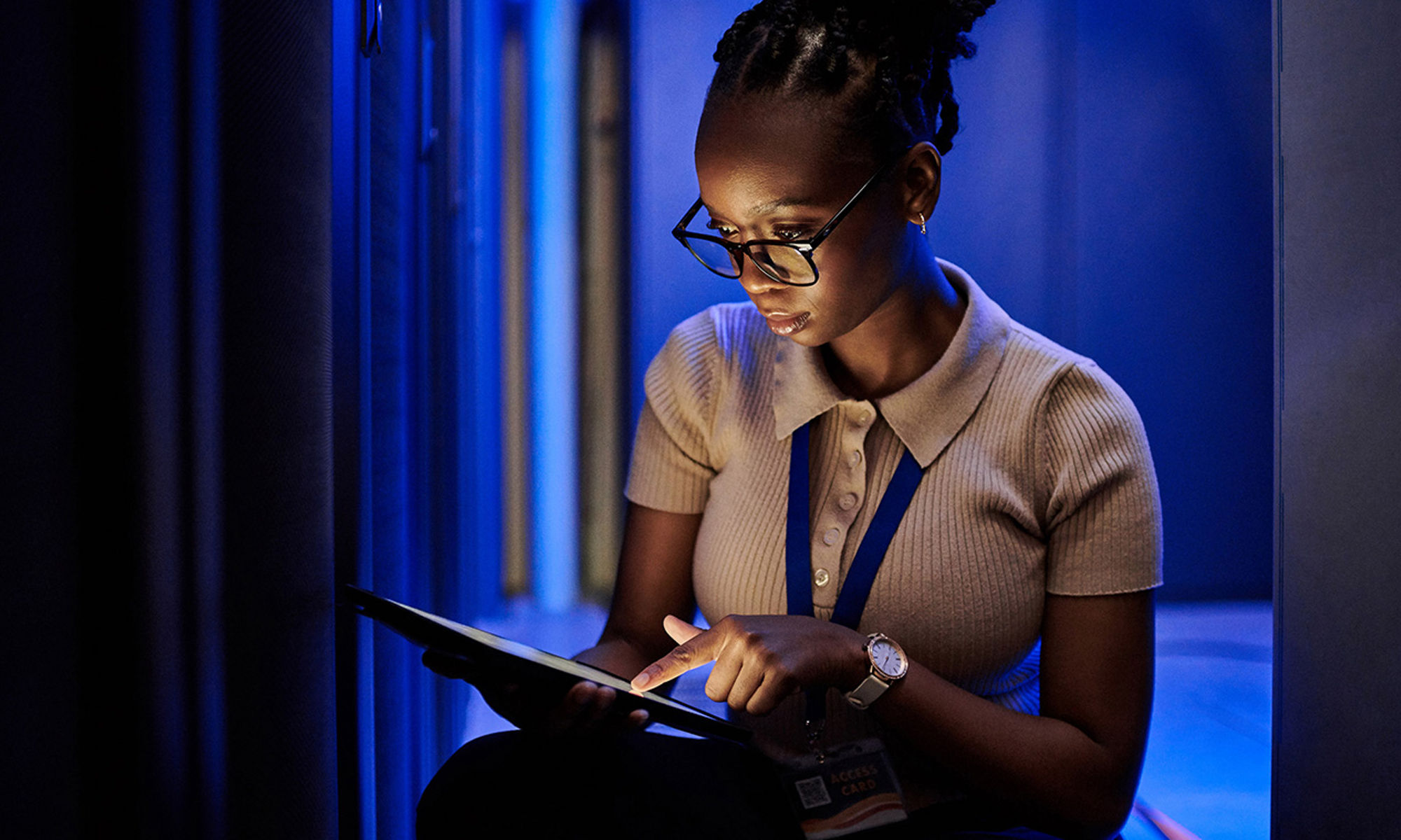 Woman kneeling, working on her laptop, in a darkened server room.