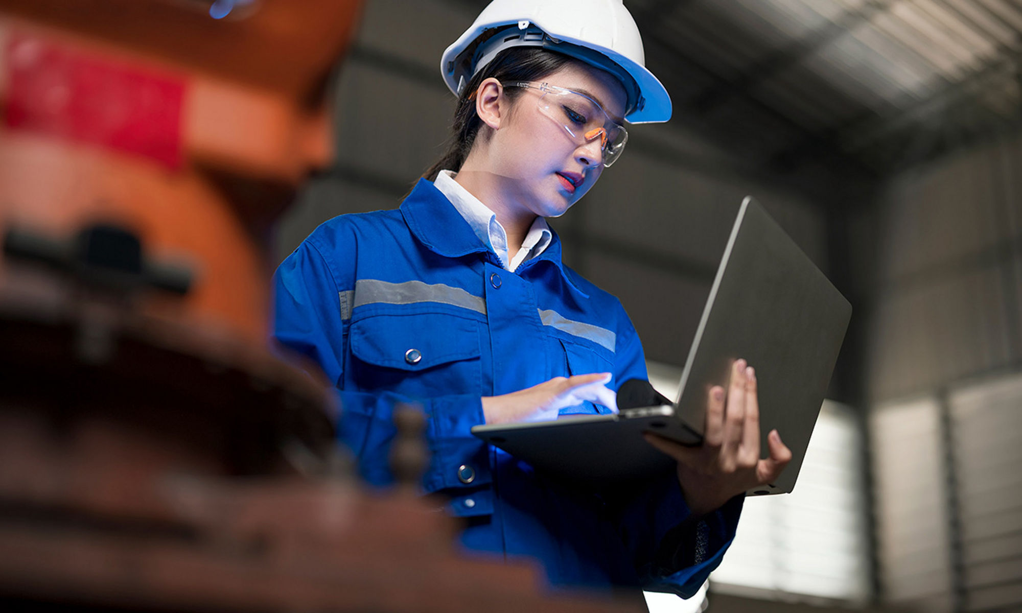 Woman worker, in a hardhad, standing up using her laptop on a manufacturing environment