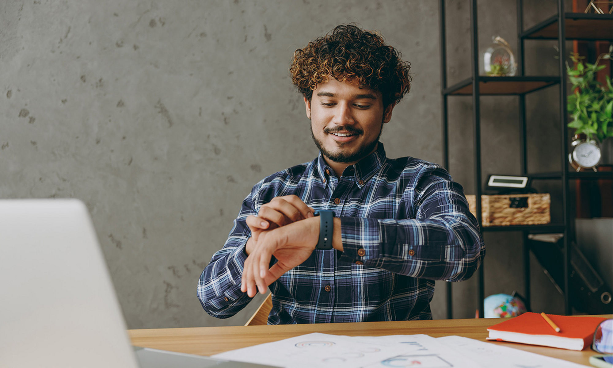 Successful smiling happy satisfied fun young employee business Indian man he wear casual blue checkered shirt looking at smart watch check time sit work at office desk with laptop pc computer indoors