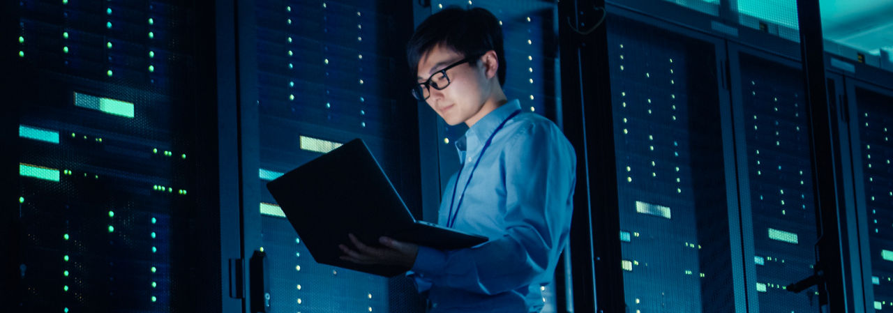 Technician with glasses using a laptop in a server room, representing Micron's contribution to fast memory and storage