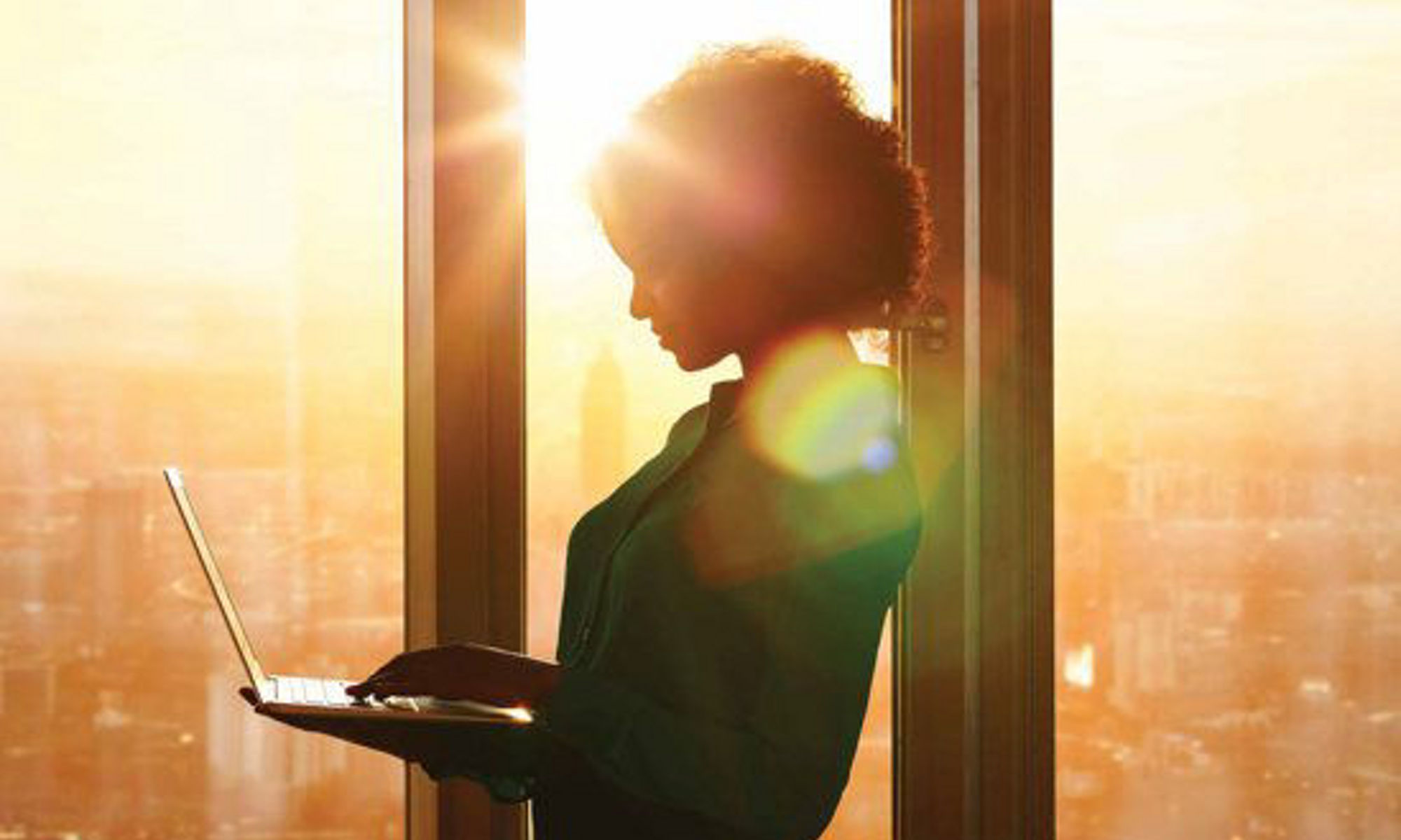 一位有色人種女性站在窗前，在明亮的陽光下與筆記型電腦互動