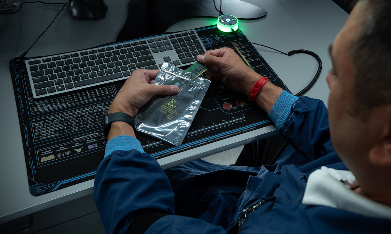 A person sitting at a desk with a computer keyboard, holding an electronic component in a static-free bag